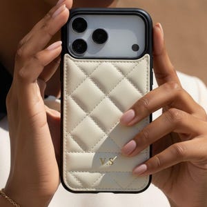 Op de afbeelding: Een zwarte en crèmekleurige telefoonhoes met een gewatteerd ontwerp. De hoes heeft een kaarthouder aan de achterkant en goudkleurige initialen "V.S.". De telefoonhoes wordt door een persoon vastgehouden.