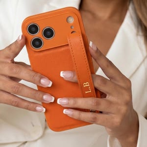 Op de afbeelding: Een feloranje smartphonehoesje met een getextureerde afwerking, voorzien van een bijpassende oranje band aan de achterkant. De band is voorzien van de goudkleurige initialen L.B. Het hoesje heeft een verhoogde uitsparing voor drie lenzen.