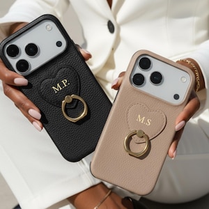 May include: Two personalized phone cases. One is black, the other is tan. Both have a heart-shaped accent with initials and a gold ring holder. The cases are designed to fit a smartphone.