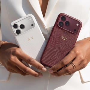 Puede incluir: Dos fundas para smartphone con un patrón texturizado de piel de lagarto, una en blanco con las iniciales A.V en dorado y otra en burdeos intenso con las iniciales V.B en dorado. Ambas fundas cuentan con un recorte para la cámara.