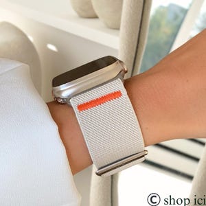 May include: A silver smartwatch with a light gray band featuring an orange stripe. The band is secured on a wrist. The watch is worn with a white long-sleeve shirt. The shop icicco logo is in the bottom right corner.