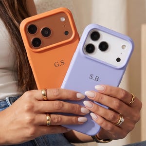 May include: Two silicone phone cases with camera cutouts. One case is orange with a brown camera frame and the initials G.S. The other case is light purple with a white camera frame and the initials S.B. Both cases feature a smooth matte finish.