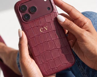 May include: A burgundy smartphone case with a textured crocodile skin pattern and the gold-colored initials C.V. embossed in the center. The case features a cutout for a triple-lens camera system and a flash, matching the deep red color scheme.