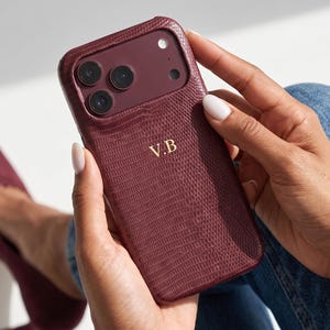 May include: A deep burgundy smartphone case with a textured lizard skin pattern and the gold embossed initials V.B. The case features a raised camera cutout section with three circular lens openings and a small circular flash opening at the top right.