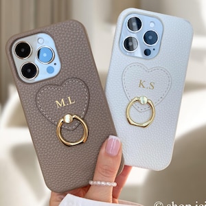 Könnte beinhalten: Zwei personalisierte Handyhüllen. Eine ist taupefarben, die andere weiß. Beide haben ein herzförmiges Design mit Initialen und einen goldenen Ringhalter. Die Hüllen bestehen aus strukturiertem Material.