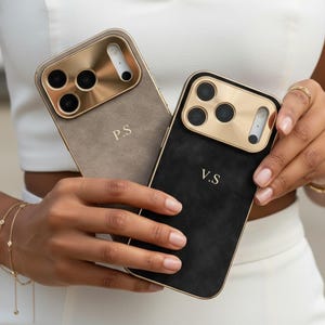 May include: Two smartphone cases with a suede-like texture, one in beige and one in black. Each case features a metallic gold camera surround and gold-toned lettering, with the beige case marked P.S and the black case marked V.S.
