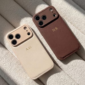以下が含まれることがあります： 質感のある仕上げの長方形のスマートフォンケース2点。1点はクリーム色で金色の文字A.Gが刻印されており、もう1点はダークブラウンで金色の文字N.Sが刻印されています。どちらも同系色の盛り上がったカメラ用カットアウトを備えています。