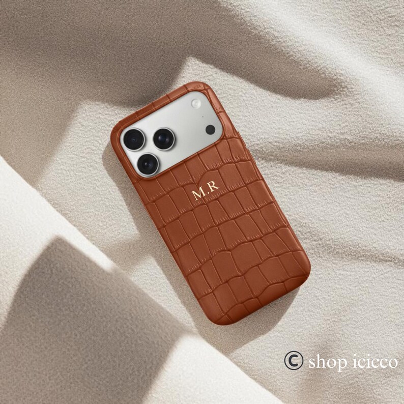 Puede incluir: Funda de tel&eacute;fono marr&oacute;n con textura de piel de cocodrilo. La funda tiene un recorte para la c&aacute;mara y las iniciales "M.R" est&aacute;n grabadas en la parte delantera. Fondo de tela de color neutro.
