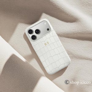 Puede incluir: Funda de tel&eacute;fono blanca con textura de piel de cocodrilo. La funda tiene un panel de c&aacute;mara plateado con tres lentes negras y una lente negra. Las iniciales "B.S" est&aacute;n impresas en dorado en la funda.