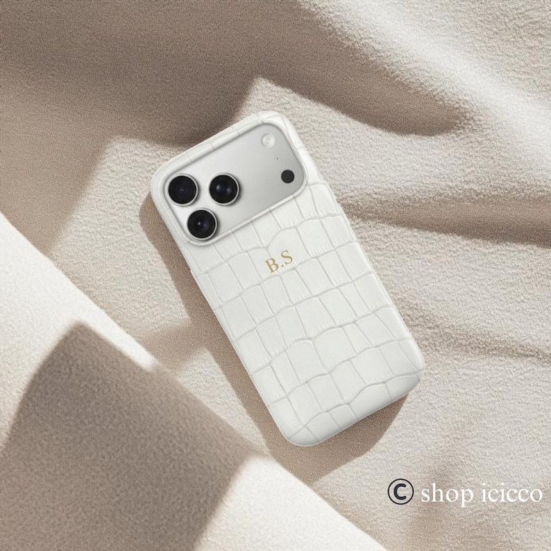 Puede incluir: Funda de tel&eacute;fono blanca con textura de piel de cocodrilo. La funda tiene un panel de c&aacute;mara plateado con tres lentes negras y una lente negra. Las iniciales "B.S" est&aacute;n impresas en dorado en la funda.