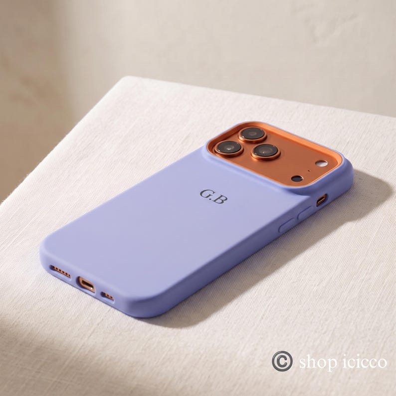 May include: A light purple smartphone case featuring a metallic copper-colored camera frame and the initials G.B printed in black on the back. The case includes precise cutouts for the camera lenses, flash, and charging port.