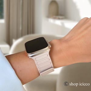 May include: A silver smartwatch with a light pink band is worn on a wrist. The watch has a rectangular screen and a metallic band with rectangular links. The watch is a modern accessory.