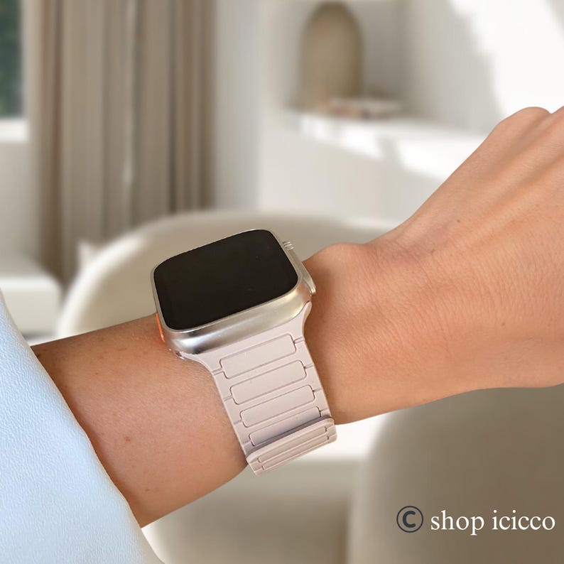 May include: A silver smartwatch with a light pink band is worn on a wrist. The watch has a rectangular screen and a metallic band with rectangular links. The watch is a modern accessory.