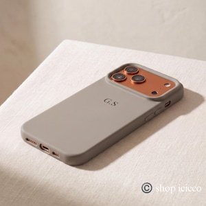 May include: A matte taupe phone case featuring a metallic copper-colored camera module with three circular lens cutouts and a small flash opening. The back of the case is personalized with the black initials G.S printed in a serif font.