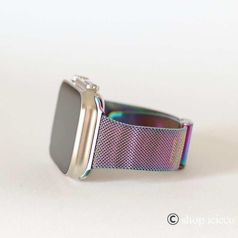 K&ouml;nnte beinhalten: Eine silberne Smartwatch mit rechteckigem Bildschirm und einem metallisch schillernden Mesh-Armband. Das Armband wechselt durch Farbt&ouml;ne von Lila, Blau und Gr&uuml;n. Die Uhr wird auf einer wei&szlig;en Oberfl&auml;che gezeigt.