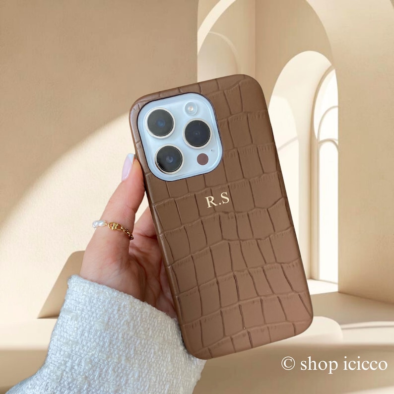Puede incluir: Funda de tel&eacute;fono marr&oacute;n con textura de piel de cocodrilo. La funda tiene un recorte para la c&aacute;mara y las iniciales "R.S" impresas en la parte delantera. La funda la sostiene una persona que lleva un su&eacute;ter blanco y un anillo dorado.