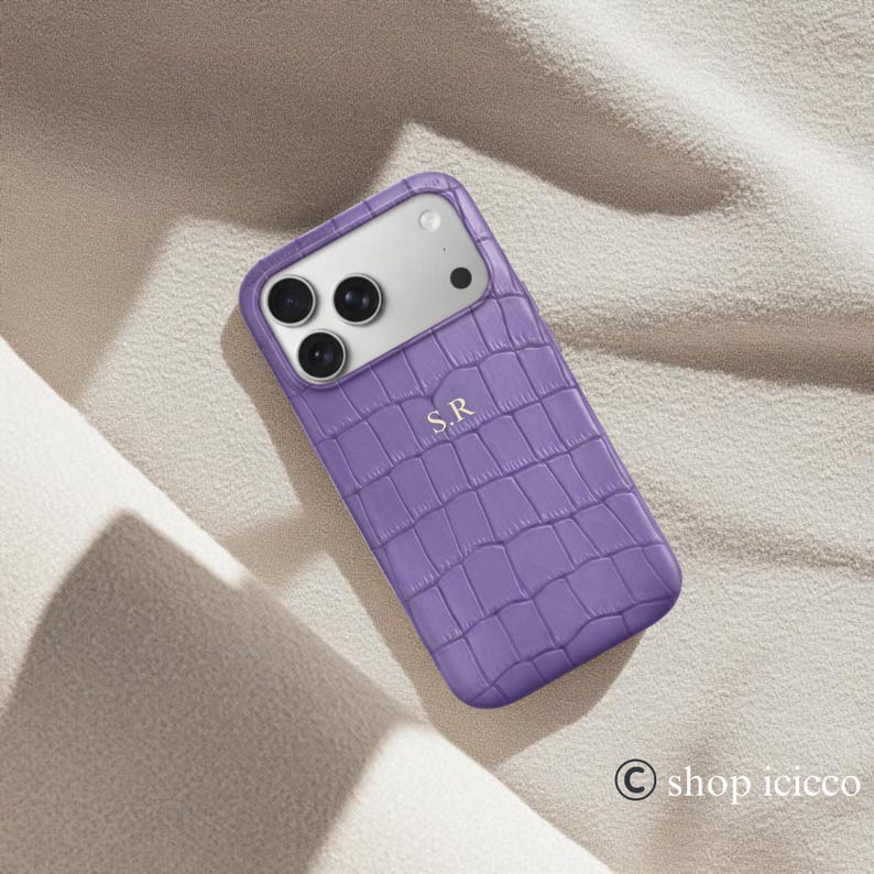Puede incluir: Una funda de tel&eacute;fono morada con textura de piel de cocodrilo. La funda tiene un m&oacute;dulo de c&aacute;mara plateado y las iniciales "S.R" en relieve en la parte delantera. La funda est&aacute; sobre una superficie clara y texturizada.
