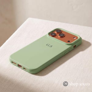 May include: A sage green smartphone case featuring a metallic copper-colored camera module with three circular lens cutouts and a flash. The back of the case is personalized with the black initials G.S. centered in a simple serif font.