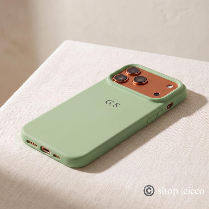 May include: A sage green smartphone case featuring a metallic copper-colored camera module with three circular lens cutouts and a flash. The back of the case is personalized with the black initials G.S. centered in a simple serif font.