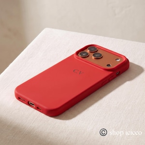 May include: A bright red smartphone case featuring a metallic copper-toned camera module surround and the initials C.V. embossed in the center. The case includes precise cutouts for the camera lenses, flash, charging port, and side buttons.