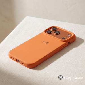 May include: A matte orange smartphone case featuring a raised camera cutout with a metallic copper-toned border. The back of the case is personalized with the black initials G.S. centered in the middle. The case includes precise cutouts for the charging port.
