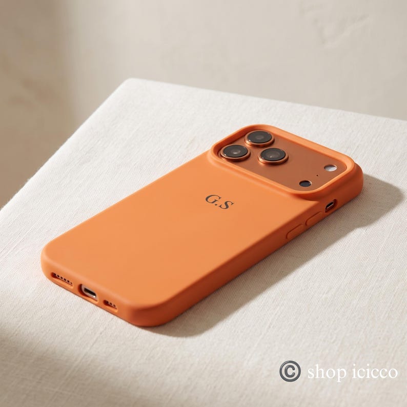 May include: A matte orange smartphone case featuring a raised camera cutout with a metallic copper-toned border. The back of the case is personalized with the black initials G.S. centered in the middle. The case includes precise cutouts for the charging port.