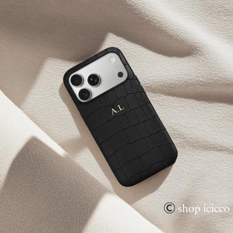 Puede incluir: Funda de tel&eacute;fono negra con textura de piel de cocodrilo. La funda tiene un m&oacute;dulo de c&aacute;mara plateado y las iniciales "A.L" en dorado. La funda est&aacute; sobre una tela beige.