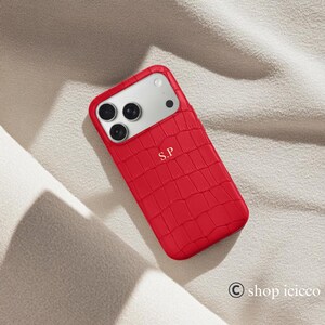 Puede incluir: Funda de tel&eacute;fono roja con un patr&oacute;n texturizado similar a la piel de cocodrilo. La funda tiene un recorte para la c&aacute;mara y las iniciales "S.P" est&aacute;n grabadas en la parte inferior. La funda est&aacute; sobre una superficie blanca.