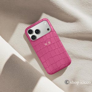 Puede incluir: Una funda de tel&eacute;fono rosa brillante con textura de piel de cocodrilo. La funda tiene una secci&oacute;n de c&aacute;mara plateada y las iniciales "W.B" son visibles. La funda est&aacute; sobre una superficie de color neutro. El s&iacute;mbolo de copyright y "shop icicco" est&aacute;n abajo a la derecha.