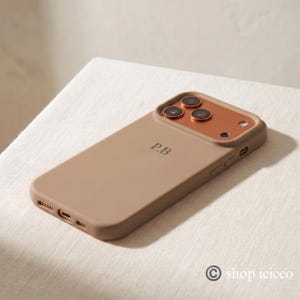 May include: A matte beige smartphone case featuring the initials P.B in a dark serif font. The case includes a metallic copper-colored camera module surround with three circular lens cutouts and a small circular flash opening on the top right.