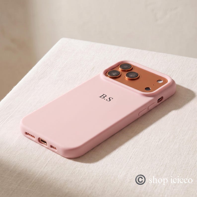 May include: A light pink smartphone case featuring a metallic copper-colored camera lens frame and the initials B.S printed in black on the back. The case includes precise cutouts for the camera lenses, flash, and charging port.