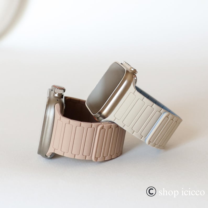 May include: Two smartwatches with bands in neutral colors. One watch has a light tan band, and the other has a pinkish-brown band. The watch faces are silver and black. The watches are on a white background.
