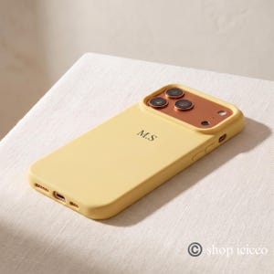 May include: A pale yellow smartphone case featuring a metallic copper-toned camera housing with three circular lens cutouts. The back of the case is personalized with the black initials M.S. centered in a serif font, with side buttons visible.