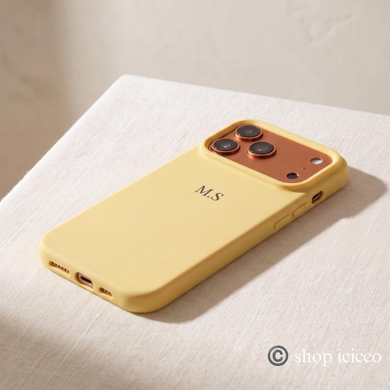 May include: A pale yellow smartphone case featuring a metallic copper-toned camera housing with three circular lens cutouts. The back of the case is personalized with the black initials M.S. centered in a serif font, with side buttons visible.