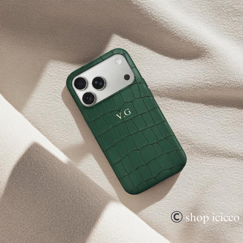Puede incluir: Funda de tel&eacute;fono verde oscuro con textura de piel de cocodrilo. La funda tiene un recorte para la c&aacute;mara y las iniciales "V.G" est&aacute;n grabadas en la funda. La funda est&aacute; sobre una tela beige.