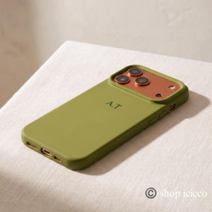 May include: An olive green smartphone case featuring a metallic copper-colored camera module and the initials A.T. engraved in the center. The case includes precise cutouts for the charging port, speakers, and side buttons, with a smooth finish.