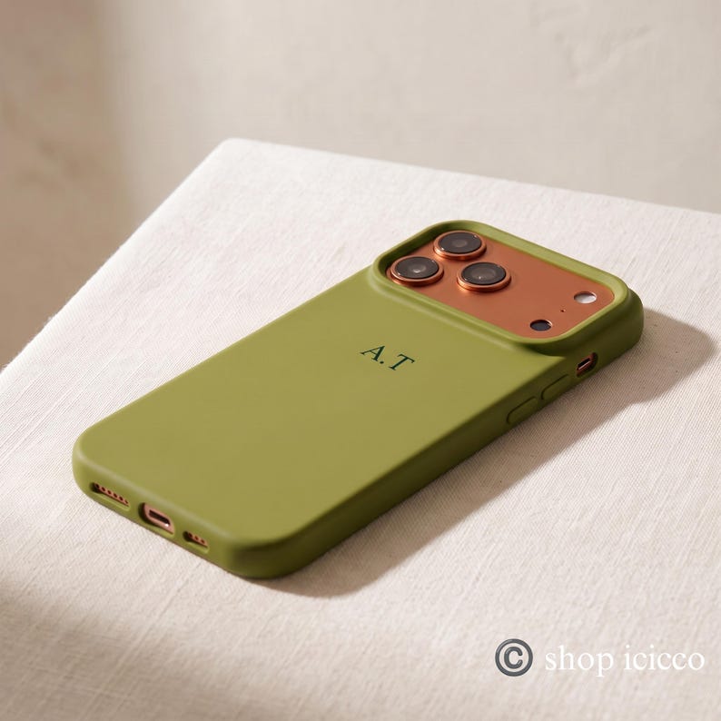 May include: An olive green smartphone case featuring a metallic copper-colored camera module and the initials A.T. engraved in the center. The case includes precise cutouts for the charging port, speakers, and side buttons, with a smooth finish.