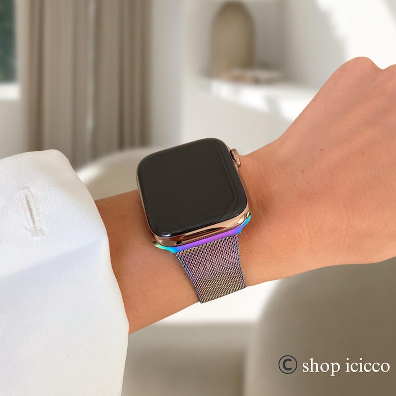 K&ouml;nnte beinhalten: Eine goldfarbene Smartwatch mit schwarzem Bildschirm und einem metallischen Armband in Regenbogenfarben wird am Handgelenk gezeigt. Das Armband hat einen Farbverlauf von Lila &uuml;ber Blau nach Gr&uuml;n. Die Uhr wird mit einem wei&szlig;en Langarmhemd getragen.