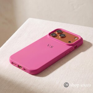 May include: A bright pink smartphone case featuring a metallic copper-colored camera module and the initials V.S printed in black on the back. The case has precise cutouts for the camera lenses, flash, and charging port, with side buttons.