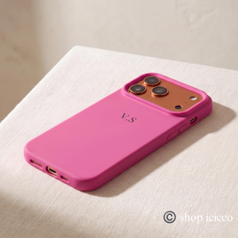 May include: A bright pink smartphone case featuring a metallic copper-colored camera module and the initials V.S printed in black on the back. The case has precise cutouts for the camera lenses, flash, and charging port, with side buttons.