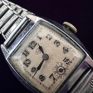 May include: A vintage rectangular wristwatch with a silver-toned metal case and an expandable link bracelet. The off-white dial features black Arabic numerals, black hands, and a small sub-dial at the six o'clock position. A gold-toned crown is on the side.