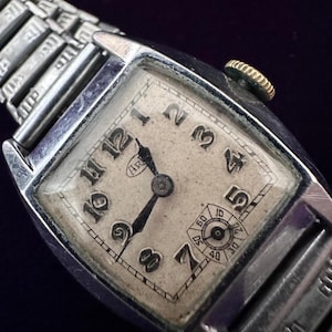 May include: A silver-toned rectangular wristwatch with a weathered off-white dial, black Arabic numerals, and black hands. The watch features a small sub-dial for seconds at the six o'clock position, a gold-toned winding crown, and a metallic expansion band.