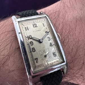 May include: A rectangular silver-tone wristwatch with a cream-colored dial featuring black Arabic numerals, the brand name Flora, and a small sub-dial for seconds. The watch has a black braided strap and the text Fab. Suisse printed at the bottom.