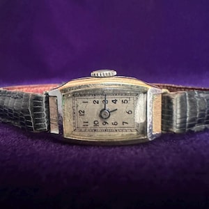 May include: A rectangular wristwatch with a silver-toned metal case and a textured black leather strap. The watch face features a white background with black Arabic numerals from 1 to 12, thin black hands, and a small winding crown at the top.