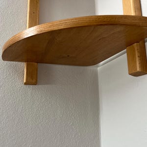 May include: A wooden corner shelf with a curved front edge. The shelf is made of light-colored wood and is supported by two vertical wooden supports. The shelf is mounted on a white wall.