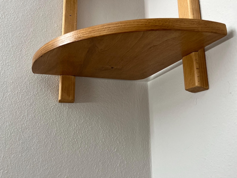 May include: A wooden corner shelf with a curved front edge. The shelf is made of light-colored wood and is supported by two vertical wooden supports. The shelf is mounted on a white wall.