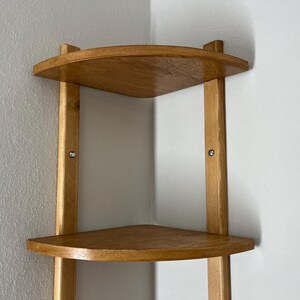 May include: A light-colored wooden corner shelf with three triangular shelves. The shelves are supported by vertical wooden posts. The shelf is designed to fit into a corner and is mounted on a white wall.