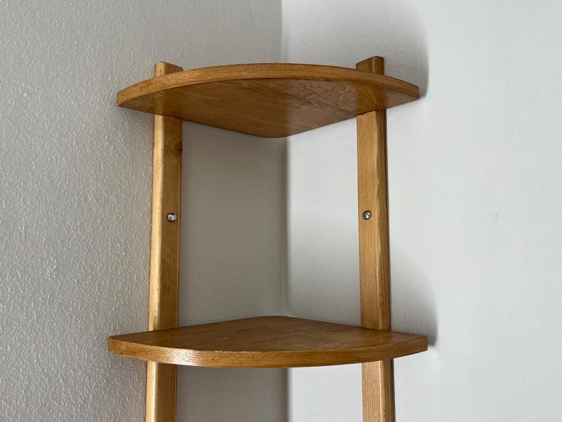 May include: A light-colored wooden corner shelf with three triangular shelves. The shelves are supported by vertical wooden posts. The shelf is designed to fit into a corner and is mounted on a white wall.