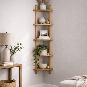 May include: A light wood corner shelf unit with five shelves, displaying decorative items. The shelves hold a white vase with tall dried grasses, ceramic pots, a framed picture, books, a potted plant, a bowl, and candles. The unit is in a neutral-toned room.