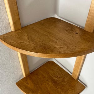May include: A two-tiered corner shelf made of light-colored wood. The shelves are triangular with rounded edges, supported by vertical and angled wooden supports. The wood grain is visible, and the shelf is mounted on a white wall.